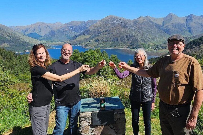 Four people standing on a grassy hill with a lake and mountains in the background. 1