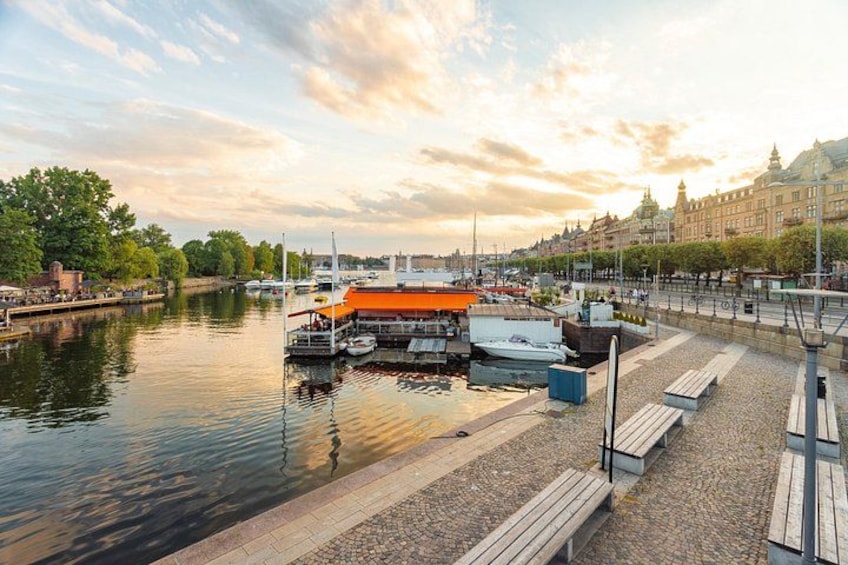 A riverside area with boats docked, a wooden walkway, and buildings in the background. 1