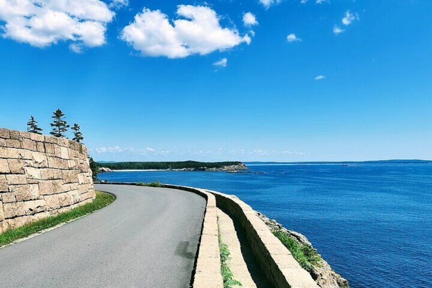 A coastal road curving alongside a body of water with a stone wall and trees on one side. 1