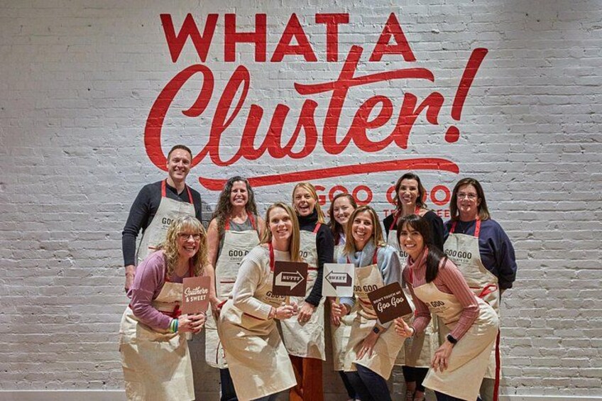 A group of people in aprons with signs, standing in front of a wall with the text 'WHAT A CLUSTER!' 2