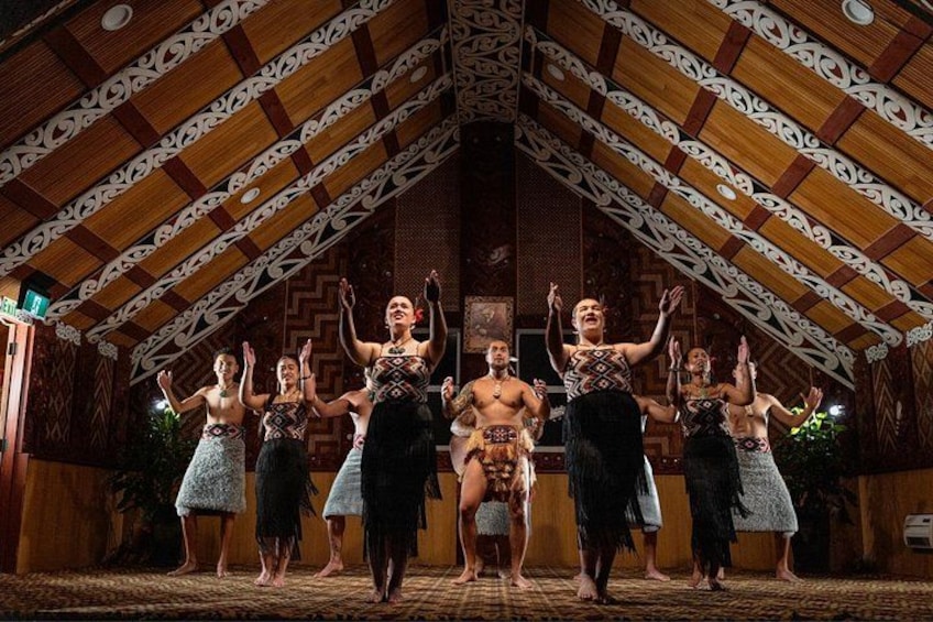 A group of dancers in traditional attire performing on a stage with a wooden ceiling and patterned walls. 1