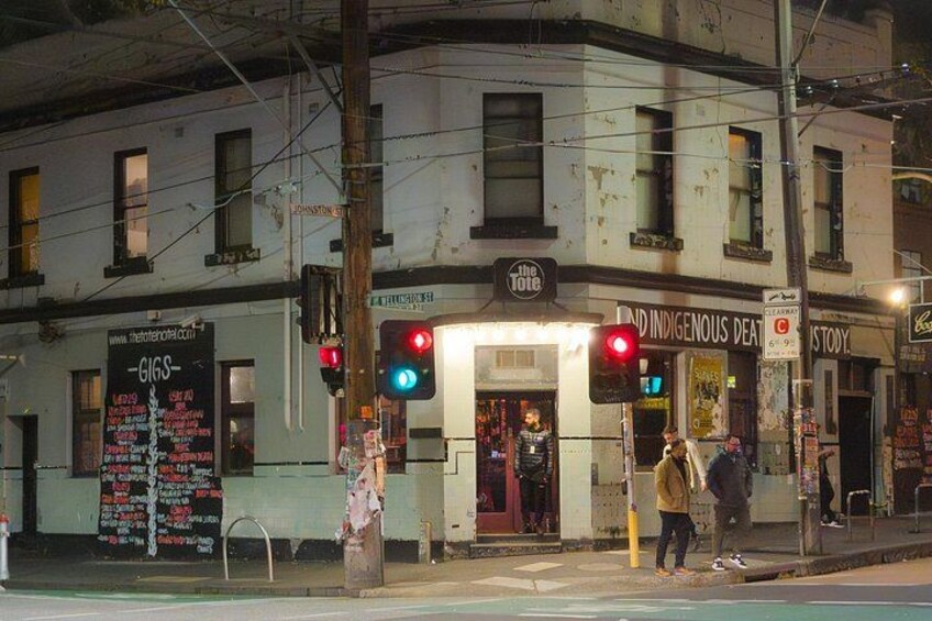 A street corner with a building that has a sign for 'The Tote' and another for 'Indigenous Dance.' There are traffic lights and a pedestrian crossing. 2