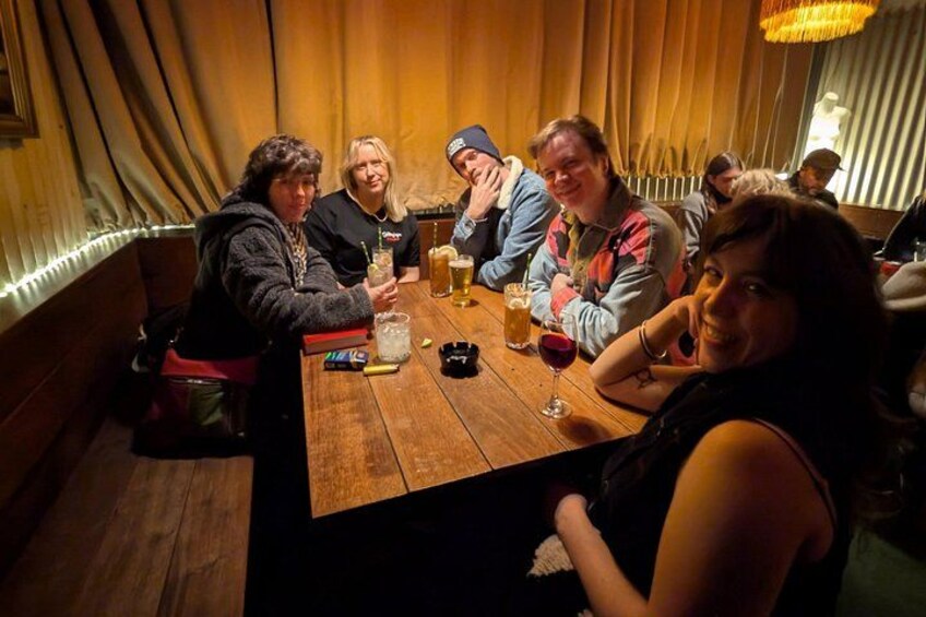 Een groep mensen zit aan een houten tafel in een schemerige kamer met gordijnen. 4