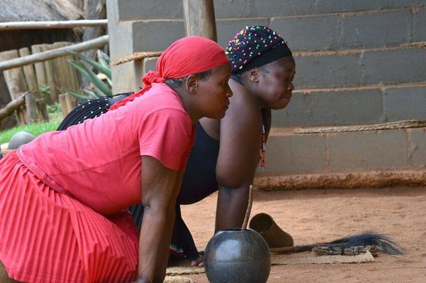 Two women in traditional attire, one in red and the other in black, sitting on the ground with a large pot in front of them. 5