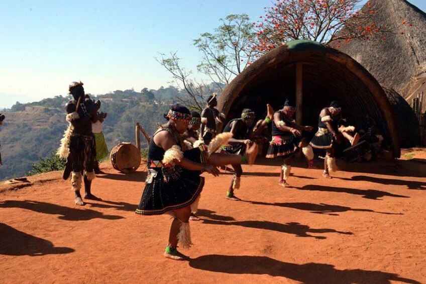 A group of people performing a traditional dance in front of a thatched hut. 1