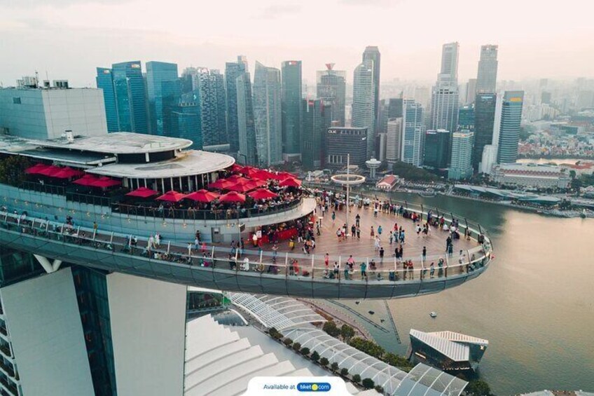 A waterfront promenade with a circular viewing platform, red umbrellas, and a city skyline in the background. 1