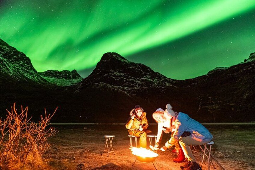 Eine Gruppe von Menschen versammelt sich an einem Lagerfeuer in einer Bergregion, im Hintergrund sind die Polarlichter zu sehen. 1