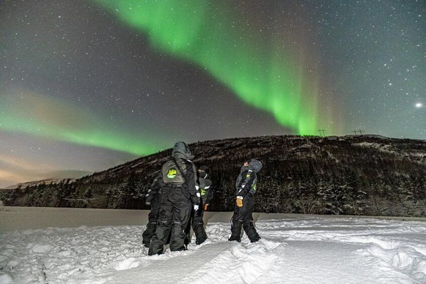 Tres personas de pie sobre terreno nevado bajo un cielo estrellado con auroras boreales. 2