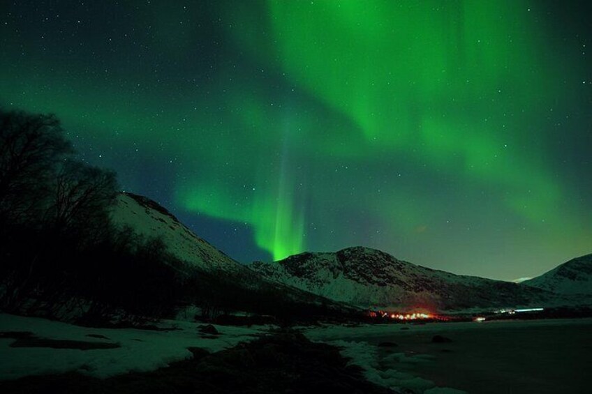 The Northern Lights over a snowy landscape with mountains and a distant light source. 2