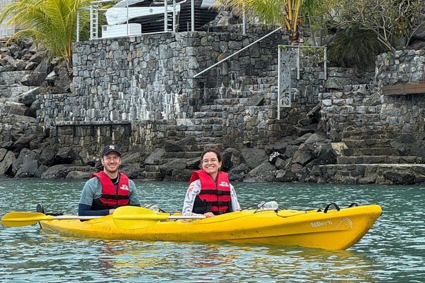 Deux personnes dans un kayak jaune sur une étendue d’eau calme, avec une côte rocheuse et des palmiers en arrière-plan. 1