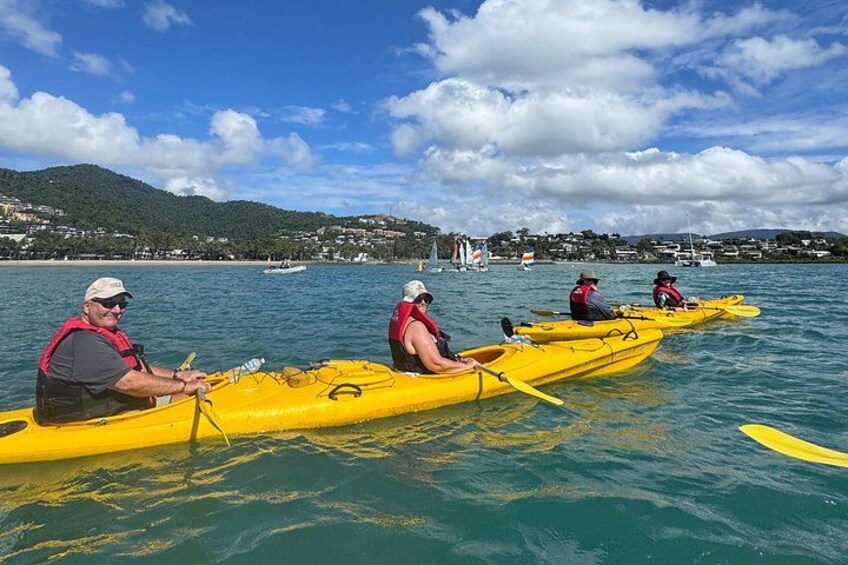 Trois personnes pratiquent le kayak sur une étendue d’eau calme, avec un paysage vallonné en arrière-plan. 4