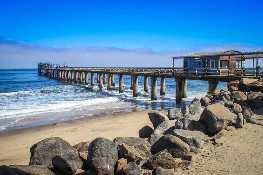 Un muelle se adentra en el océano con un edificio al final, rodeado de grandes rocas y una playa de arena. 4