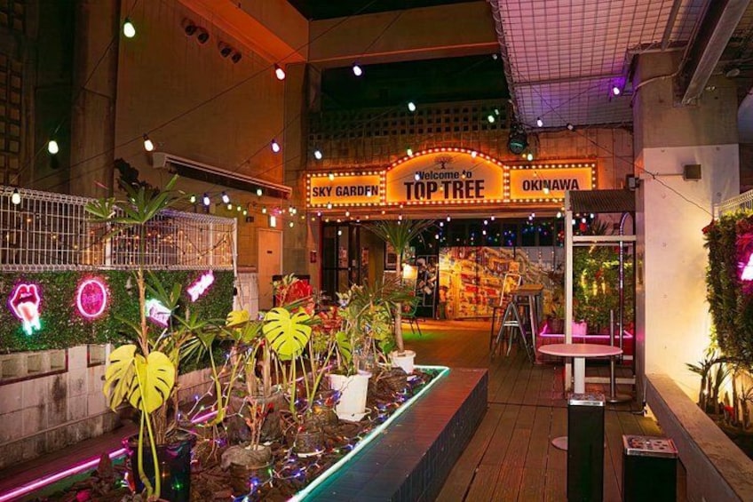 Ein hell erleuchteter Außenbereich mit einem Neon-Schild, das 'Welcome to Top Tree' und 'OKINAWA' zeigt. 4