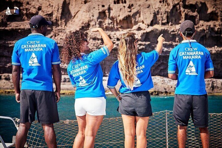 Quatre personnes portant des chemises bleues avec l’inscription « CREW CATAMARAN MAKAIARA » imprimée dans le dos, se tiennent sur un quai. En arrière-plan, on aperçoit une falaise rocheuse et l’eau. 5