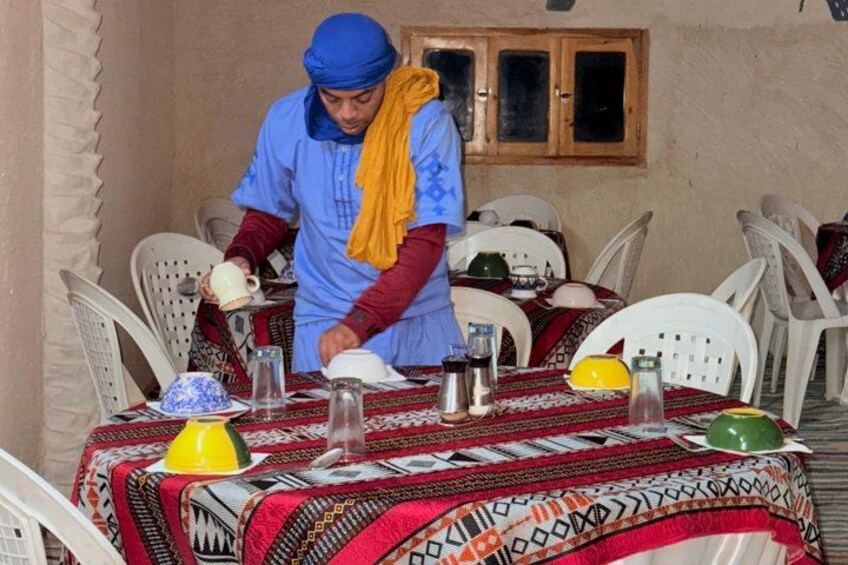 A person serving food at a table with a patterned tablecloth, white plastic chairs, and a window with wooden shutters in the background. 2