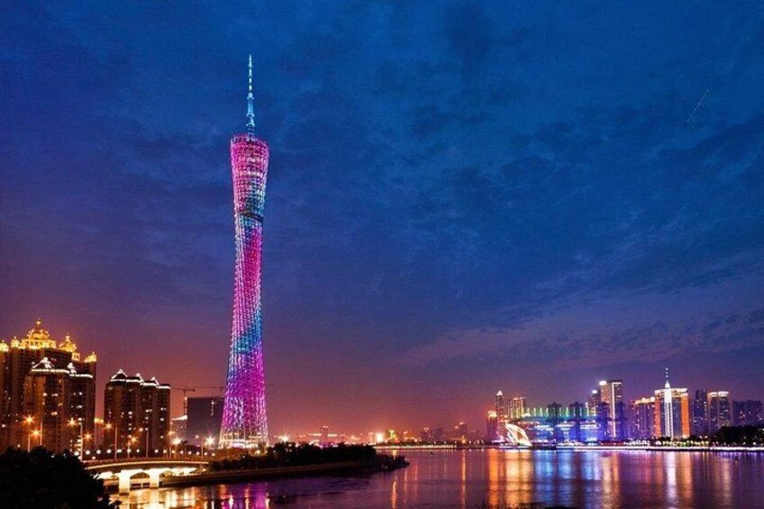 A brightly lit tower with a reflection in the water, surrounded by city buildings and a bridge. 4