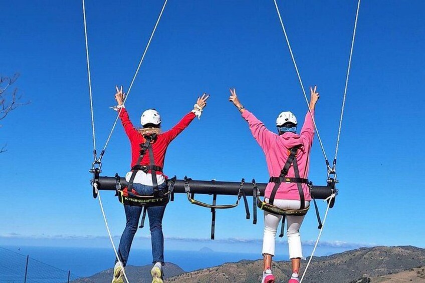 Two people on a cable car with their arms raised, overlooking a mountainous landscape and a body of water. 4