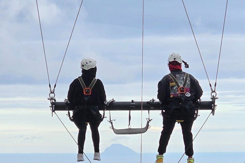 Two individuals on a cable car, wearing helmets and harnesses, with a mountainous landscape in the background. 2