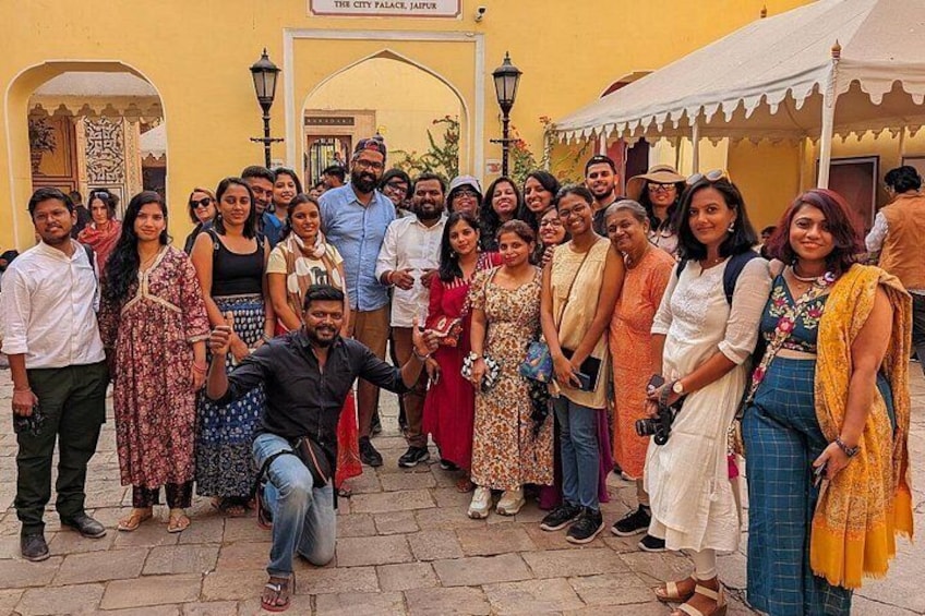 A group of people posing in front of a building with arches and a sign that reads 'The City Palace, Jaipur.' 1