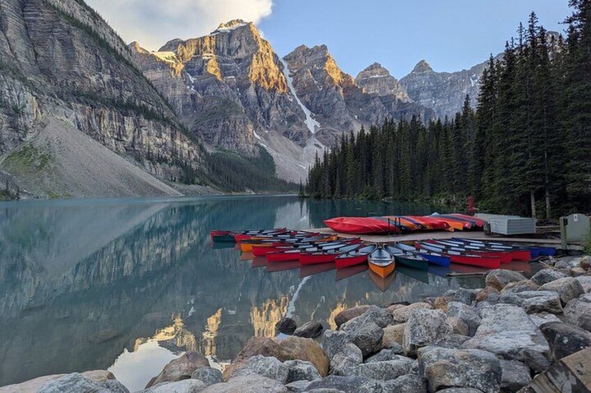 A calm lake with kayaks lined up on the shore, surrounded by mountains and trees. 1