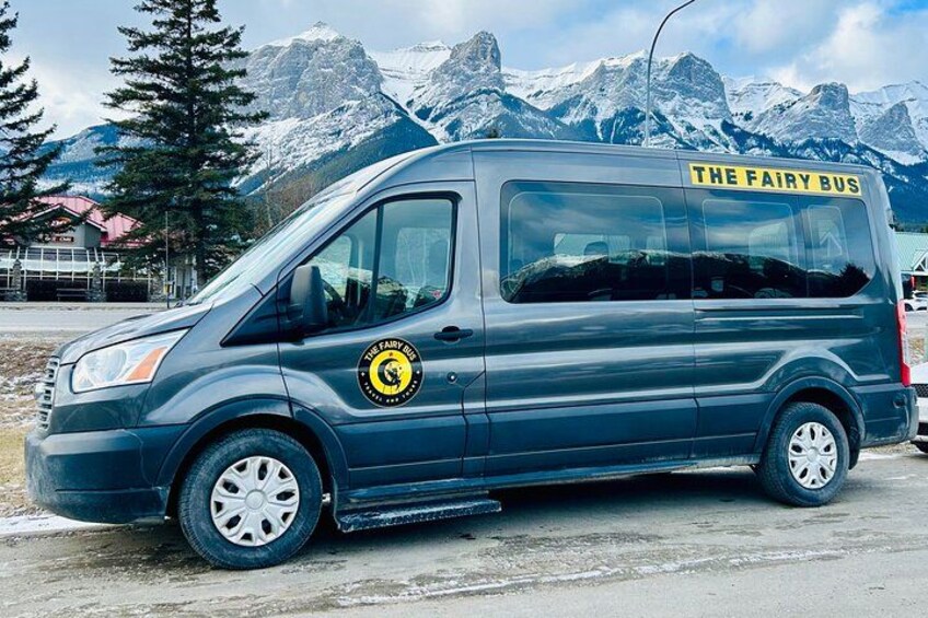 A dark blue van with 'The Fairy Bus' written on the side, parked on a road with snow-capped mountains in the background. 2