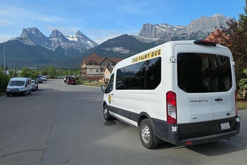 A white van with 'THE FAIRY BUS' written on the back, parked on a street with houses and mountains in the background. 4