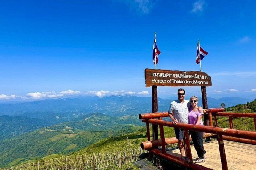 Un letrero de madera con banderas, una pareja de pie en una plataforma de madera y un paisaje montañoso. 4