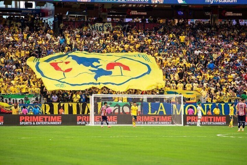 Un estadio lleno de espectadores vistiendo amarillo y azul, un gran cartel amarillo con un logotipo, y anuncios para 'PRIMETIME SPORTS & ENTERTAINMENT'. 1