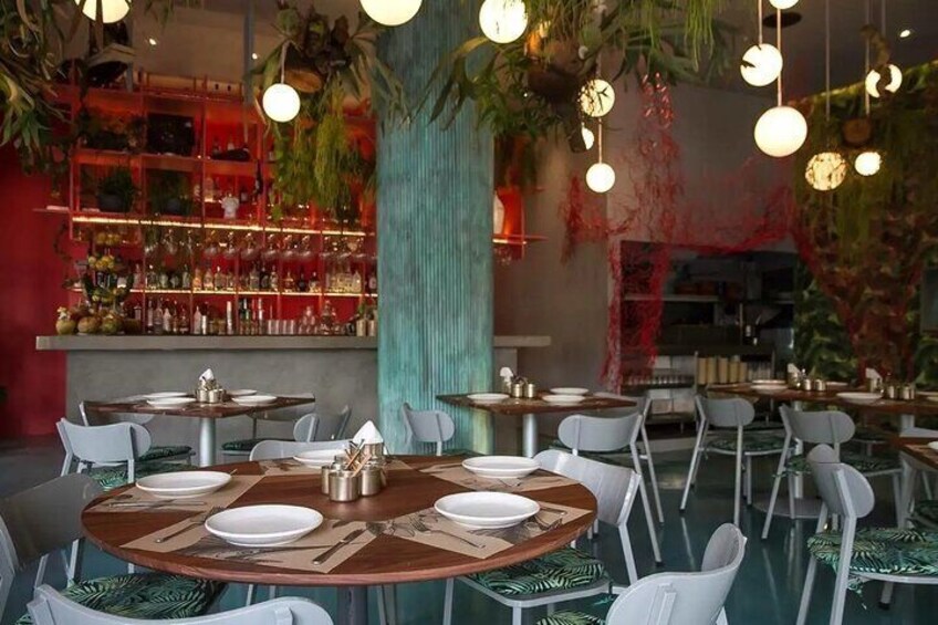 Un interior de restaurante con mesas redondas, sillas blancas, plantas colgantes y una zona de bar con estantes llenos de botellas. 5