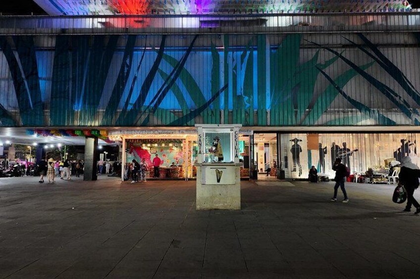 A nighttime urban square with a modern building facade, a kiosk, and people walking. 3
