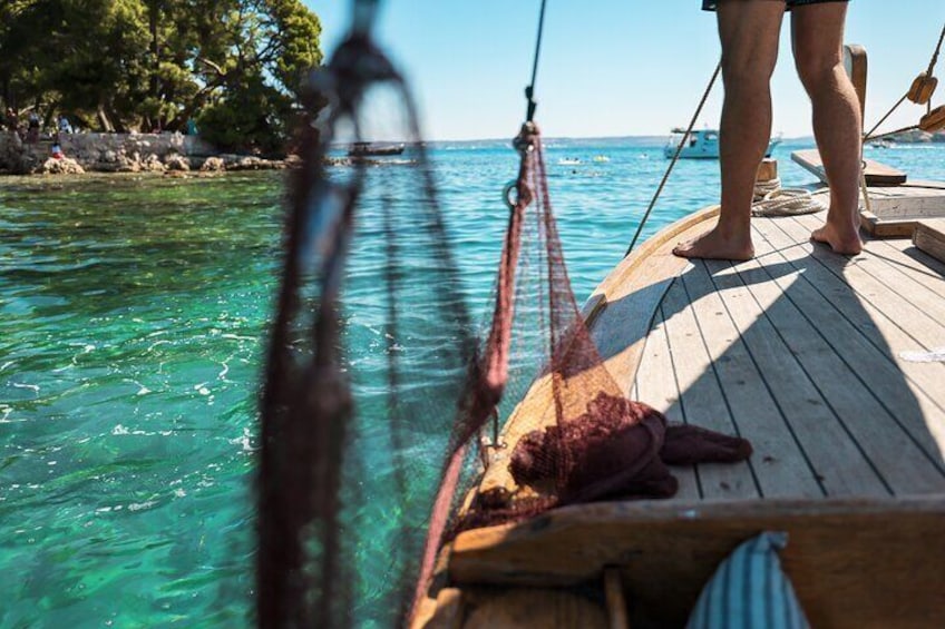 Houten dek van een boot met touwen en een net, helder turquoise water en een rotsachtige kustlijn met bomen. 5