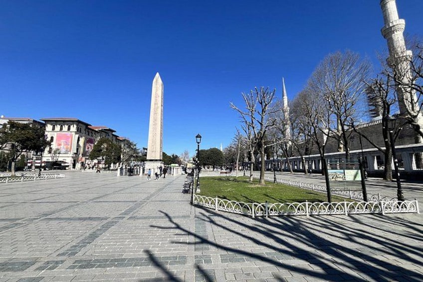 Una vasta piazza con un alto monumento, una moschea con un minareto e una recinzione alberata. 5