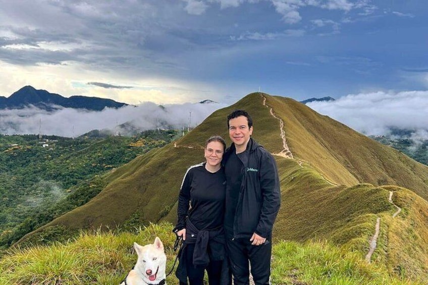 Una pareja está de pie en una colina cubierta de hierba, con un perro, contemplando un paisaje montañoso con un sendero serpenteante. 3