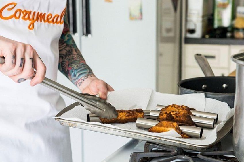 Een persoon in een witte schort met de tekst 'Cozymmeal', die met een tang eten op een grill bereidt. 2