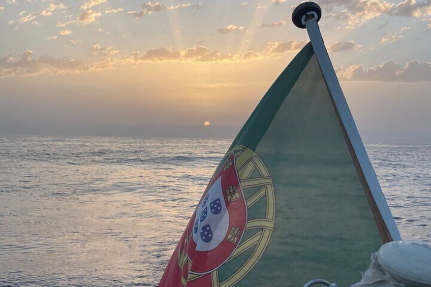 En båt med en flagga som visar ett vapensköldsmotiv, mot en solnedgång över havet. 4
