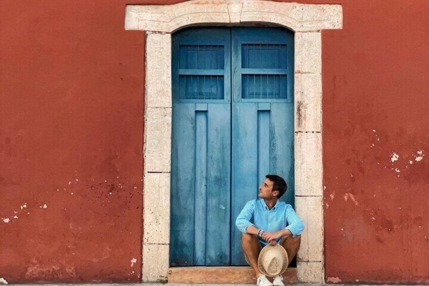A man sitting on a step in front of a blue door, wearing a light blue shirt and shorts, with a hat on the ground beside him. 4