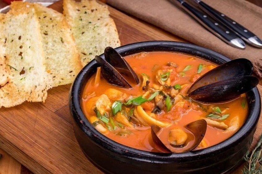 A bowl of seafood stew with mussels, accompanied by slices of toasted bread on a wooden board. 2