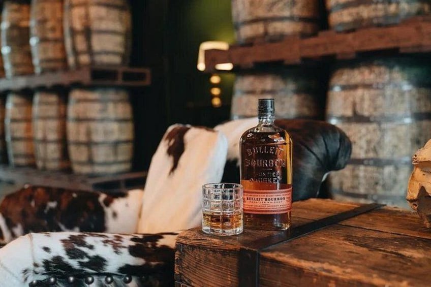 Une bouteille de whisky Bulleit Bourbon, un verre et une selle en cuir de vache posés sur une table en bois, dans un décor rustique. 4
