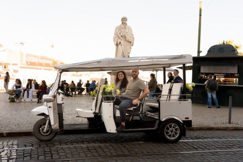 A white three-wheeled vehicle with two passengers, a statue in the background, and a crowd of people. 5