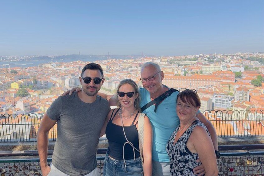A group of four people standing on a balcony overlooking a cityscape with buildings and a clear sky. 2