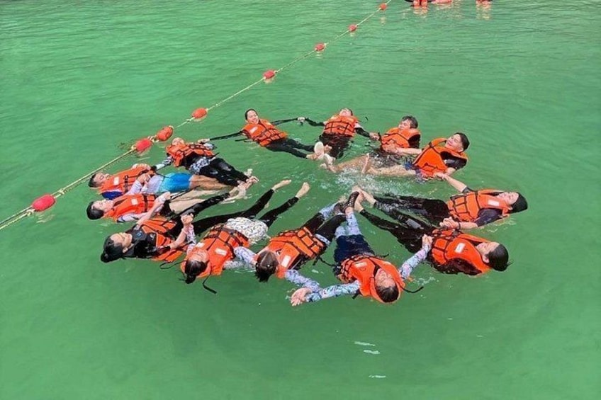 A group of people in a circle, wearing life jackets, floating on a green body of water. 2