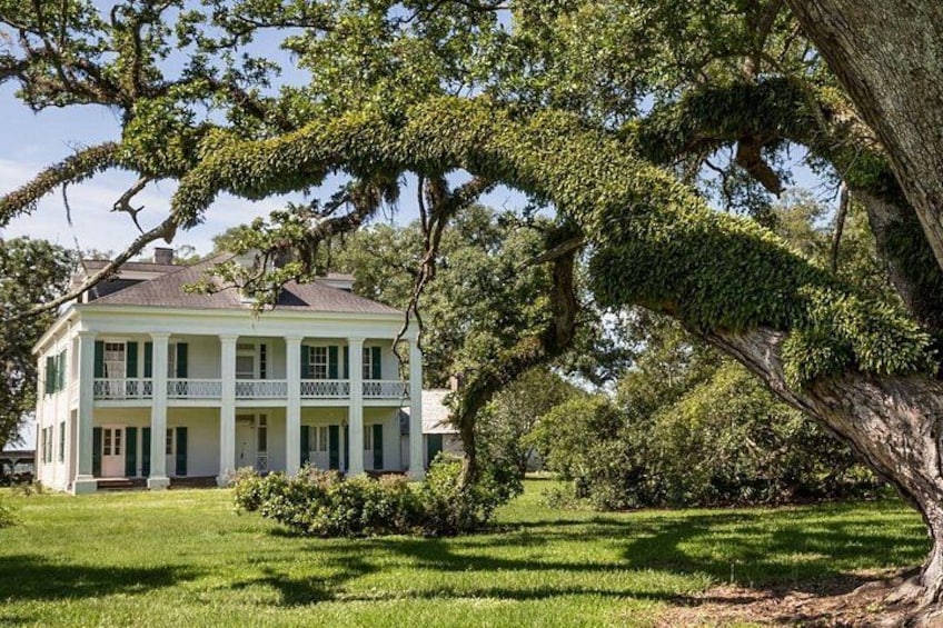 A large, white colonial-style house with a balcony, surrounded by lush greenery and trees. 1