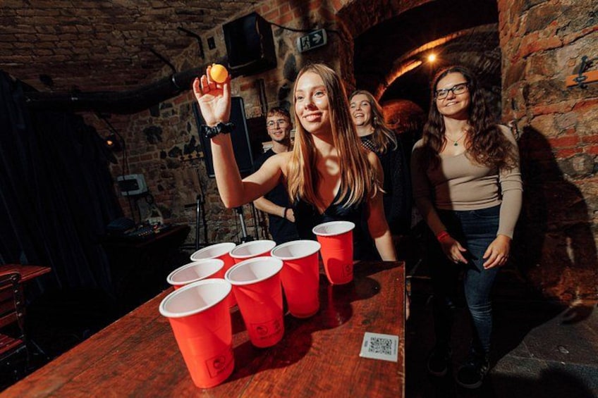 A person holding a ping pong ball above red plastic cups on a table, with a brick wall and a door in the background. 3