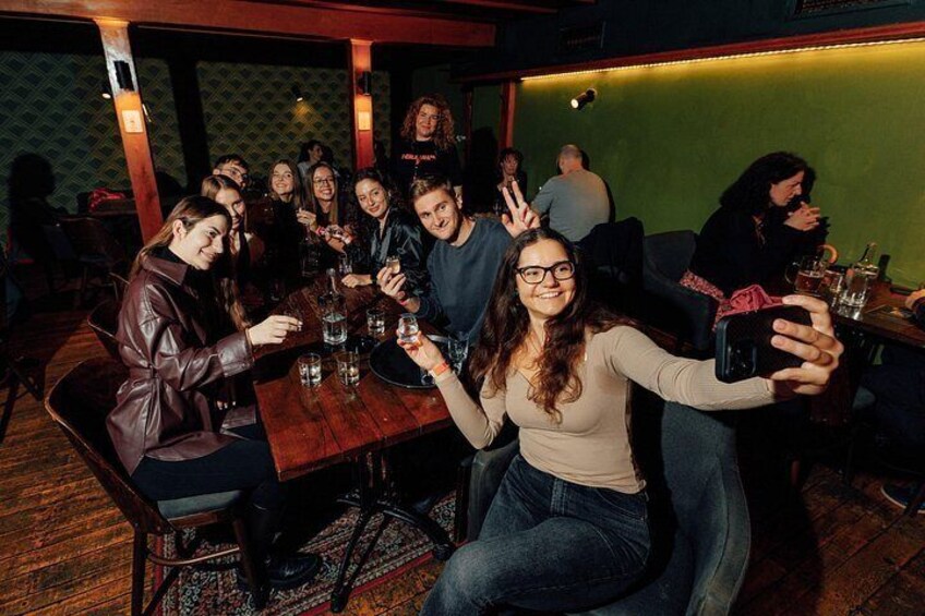 Een groep mensen zit aan een houten tafel in een kamer met gedimd licht en behang met een patroon. 3