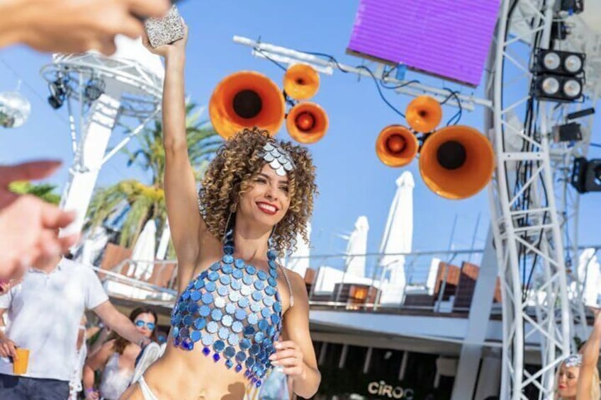 Een persoon in een blauwe paillettenoutfit met hoofdtooi, staand voor een podium met grote oranje luidsprekers. 3