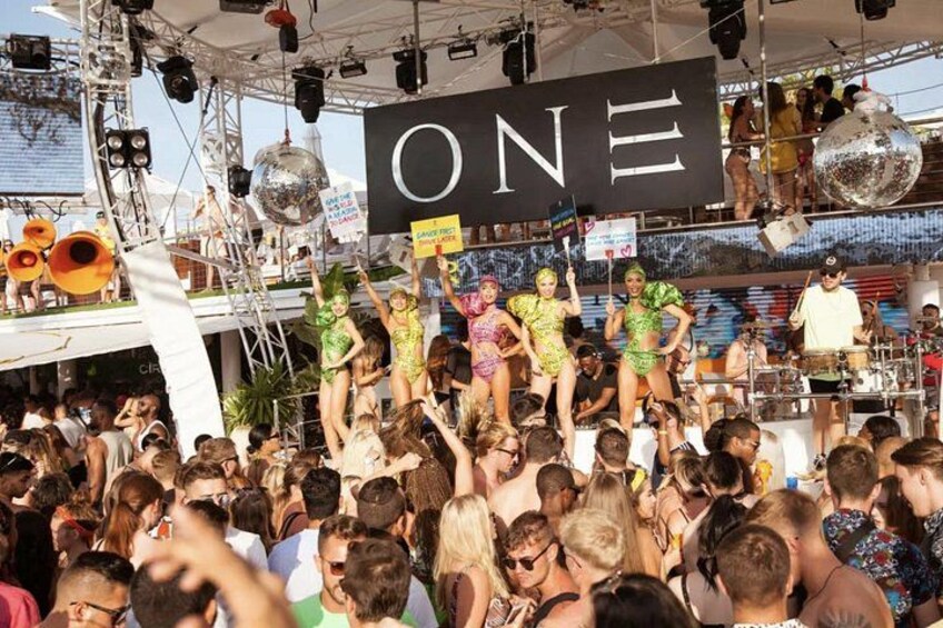 A crowded outdoor event with a stage, a large 'ONE' sign, and a disco ball. 5