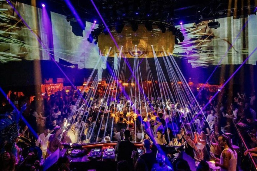 A crowded nightclub with a DJ booth, laser lights, and a large screen displaying visuals. 2