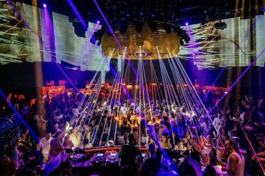 Een drukke nachtclub met een DJ-hok, laserlichten en een groot scherm waarop abstracte beelden te zien zijn. 4