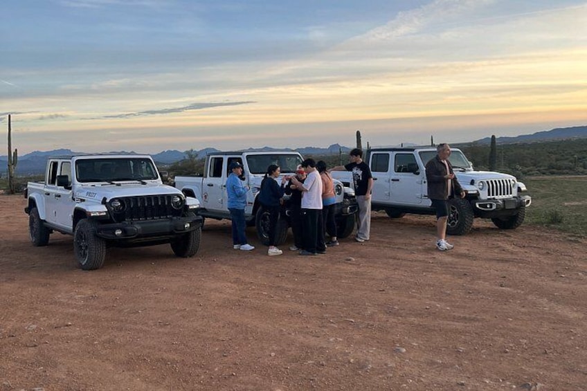 A group of people standing on a dirt road with two white pickup trucks parked nearby. 2