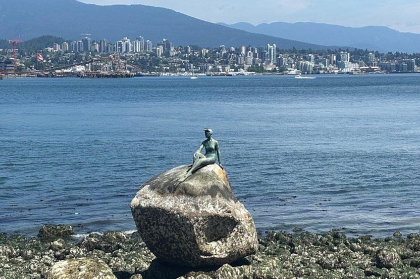 Eine Statue einer Figur auf einem Felsen an einem Gewässer, im Hintergrund eine Stadtansicht. 2